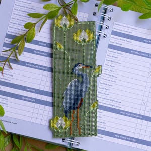 May include: A handmade bookmark featuring a cross-stitched heron design. The bookmark has a green background with yellow and white floral accents. The heron is depicted in shades of blue and gray, with orange legs. The bookmark is placed on an open journal.