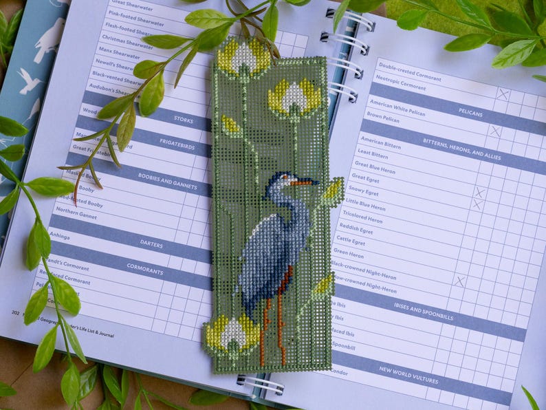 May include: A handmade bookmark featuring a cross-stitched heron design. The bookmark has a green background with yellow and white floral accents. The heron is depicted in shades of blue and gray, with orange legs. The bookmark is placed on an open journal.