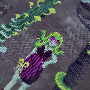 May include: Cross-stitch design featuring a Medusa-like figure with green hair and snakes, wearing a purple dress. The design includes green vines and a dark background, creating a spooky, fantasy theme. The cross-stitch is detailed and colorful.