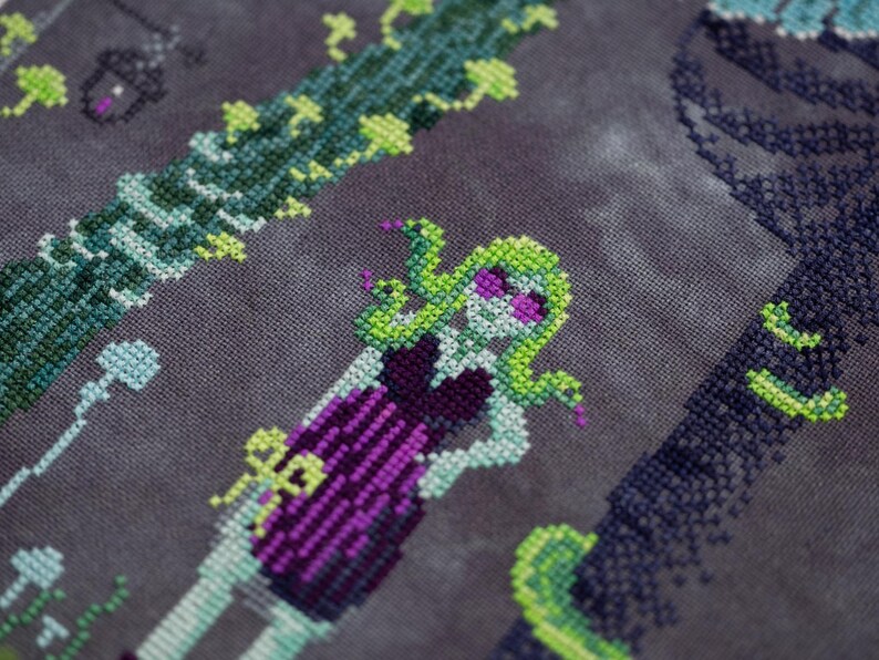 May include: Cross-stitch design featuring a Medusa-like figure with green hair and snakes, wearing a purple dress. The design includes green vines and a dark background, creating a spooky, fantasy theme. The cross-stitch is detailed and colorful.
