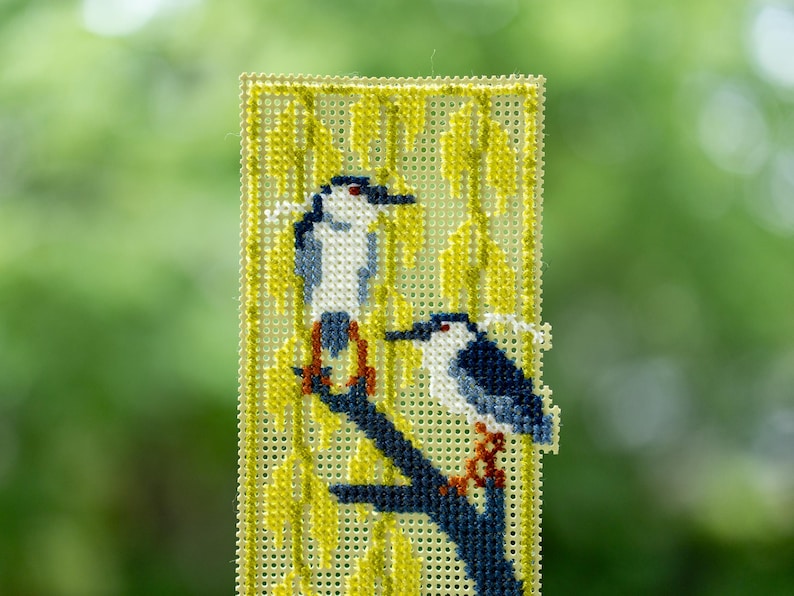 May include: A rectangular cross-stitch bookmark on perforated plastic canvas featuring two black-crowned night herons perched on a dark blue branch. The background consists of yellow willow leaves stitched in a vertical pattern against the grid.