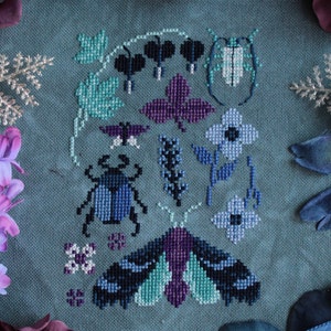 May include: A cross-stitch embroidery design featuring a blue and green moth, a beetle, and flowers on a blue fabric background.