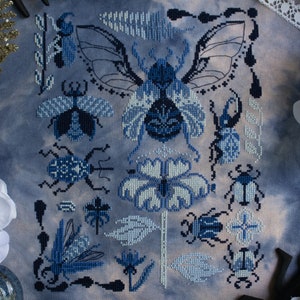 May include: A blue and white cross stitch embroidery featuring a variety of insects, including beetles, butterflies, and moths. The design is set against a blue background with white accents.