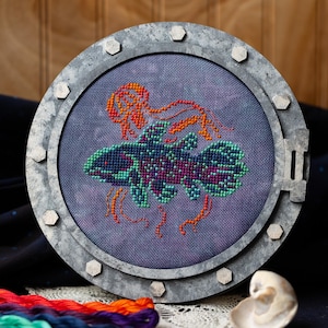 May include: Cross-stitch art of a fish and jellyfish in a round, metal-framed design. The fish is blue, green, and purple, while the jellyfish is orange. The frame has a bolted design. The background is a dark blue fabric.