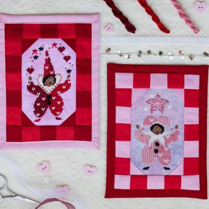 Peut inclure: Deux œuvres d'art encadrées au point de croix représentant un personnage de type clown en tenue rouge et rose. Les cadres ont un motif à carreaux rouges et roses. Les personnages sont entourés de cœurs et d'étoiles. Le fond comprend des ciseaux, un ruban et des éléments décoratifs.