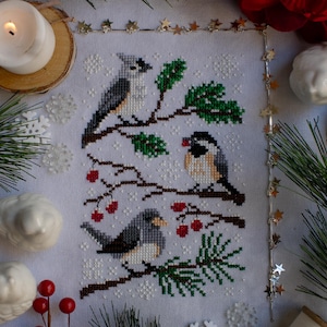 May include: Cross-stitch pattern featuring three birds perched on a snowy branch with red berries. The birds are a chickadee, a tufted titmouse, and a cardinal. The background is white with snowflakes.