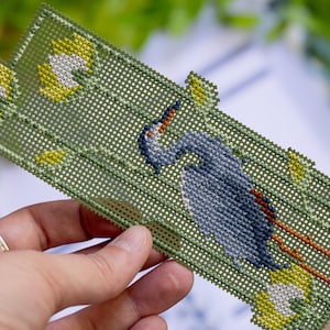 May include: Handmade needlepoint bookmark featuring a blue heron design. The bookmark has a green background with yellow and white floral accents. The heron is depicted in shades of blue and gray, with orange accents.