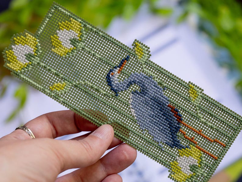 May include: Handmade needlepoint bookmark featuring a blue heron design. The bookmark has a green background with yellow and white floral accents. The heron is depicted in shades of blue and gray, with orange accents.