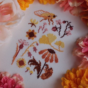Peut inclure: Broderie au point de croix repr&eacute;sentant une vari&eacute;t&eacute; de flore et de faune automnales, notamment des tournesols, des feuilles, des baies et une abeille. Le motif est brod&eacute; dans des tons de jaune, orange, marron et rose sur un fond blanc.