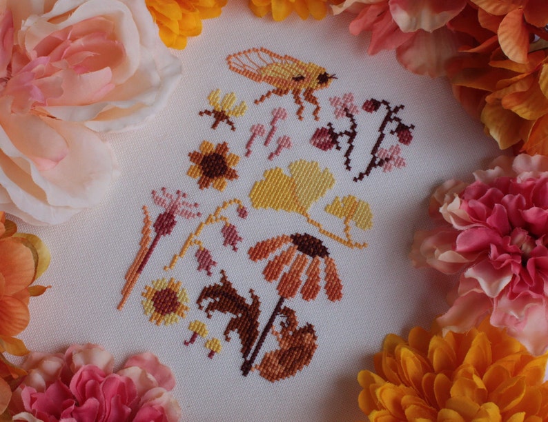 Peut inclure: Broderie au point de croix repr&eacute;sentant une vari&eacute;t&eacute; de flore et de faune automnales, notamment des tournesols, des feuilles, des baies et une abeille. Le motif est brod&eacute; dans des tons de jaune, orange, marron et rose sur un fond blanc.