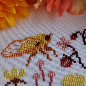 Peut inclure: Broderie au point de croix repr&eacute;sentant une abeille jaune et orange avec des ailes, entour&eacute;e de fleurs et de feuilles roses et brunes.