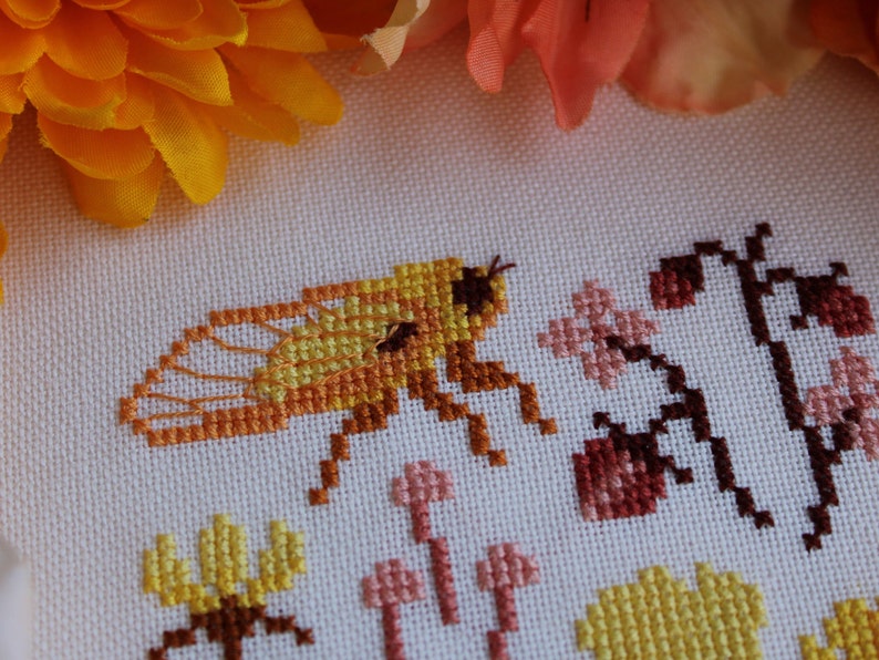 Peut inclure: Broderie au point de croix repr&eacute;sentant une abeille jaune et orange avec des ailes, entour&eacute;e de fleurs et de feuilles roses et brunes.