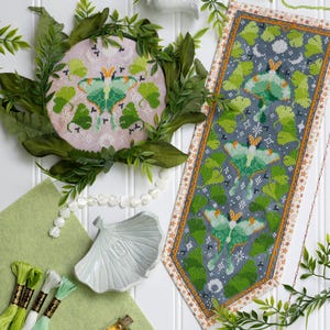 May include: A circular embroidery hoop featuring a green moth and ginkgo leaves on light pink fabric, alongside a long rectangular banner with three green moths and ginkgo leaves on dark grey fabric, accompanied by green felt and embroidery floss.