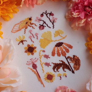 Peut inclure: Motif de point de croix avec une vari&eacute;t&eacute; de motifs floraux et d'insectes dans des tons d'orange, jaune et marron. Le motif comprend une cigale, des tournesols et d'autres fleurs sauvages.