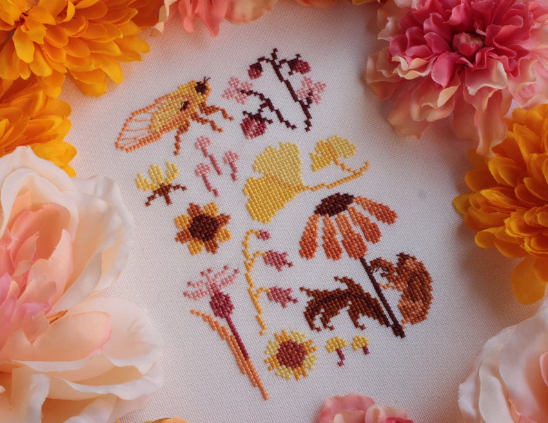 Peut inclure: Motif de point de croix avec une vari&eacute;t&eacute; de motifs floraux et d'insectes dans des tons d'orange, jaune et marron. Le motif comprend une cigale, des tournesols et d'autres fleurs sauvages.