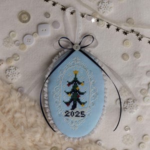 May include: A light blue, oval-shaped Christmas ornament with a cross-stitched Christmas tree design. The ornament has a white lace trim, a dark blue ribbon, and the year "2025" stitched on it. White buttons and snowflakes are scattered around the ornament.