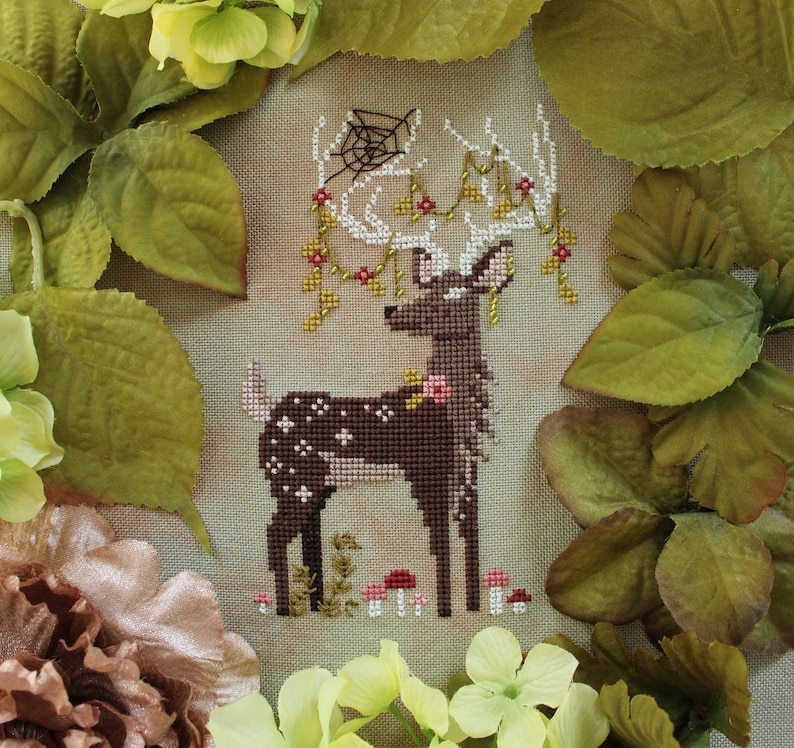 May include: A cross-stitch design of a deer with white antlers adorned with flowers and vines. The deer is brown with white spots and is surrounded by green leaves and flowers. The design includes a spiderweb.