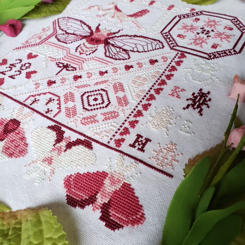 Another view of the finished pink cross stitch sampler of several insects and heart motifs, including a large cicada, praying mantis, and several moths. The initials K L H are stitched next to the beetle motifs.