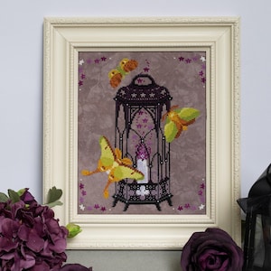 Peut inclure: Une broderie au point de croix encadrée représentant une lanterne noire avec une bougie violette allumée à l'intérieur, entourée de trois papillons jaunes et verts sur un fond mauve chiné, dans un cadre rectangulaire blanc cassé.