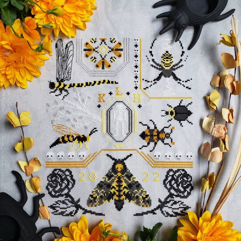 A finished cross stitch featuring two large rose motifs flanking a Deaths-head hawkmoth, a large wasp, a dragonfly, beetles, and an orb weaver spider. A simple skeleton is in the middle of the design and various skull motifs are throughout.