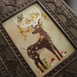 May include: Cross-stitch art of a deer with antlers adorned with a spiderweb, flowers, and berries. The deer is brown with white spots and is surrounded by mushrooms and foliage. The artwork is framed in a dark, intricately carved wooden frame.