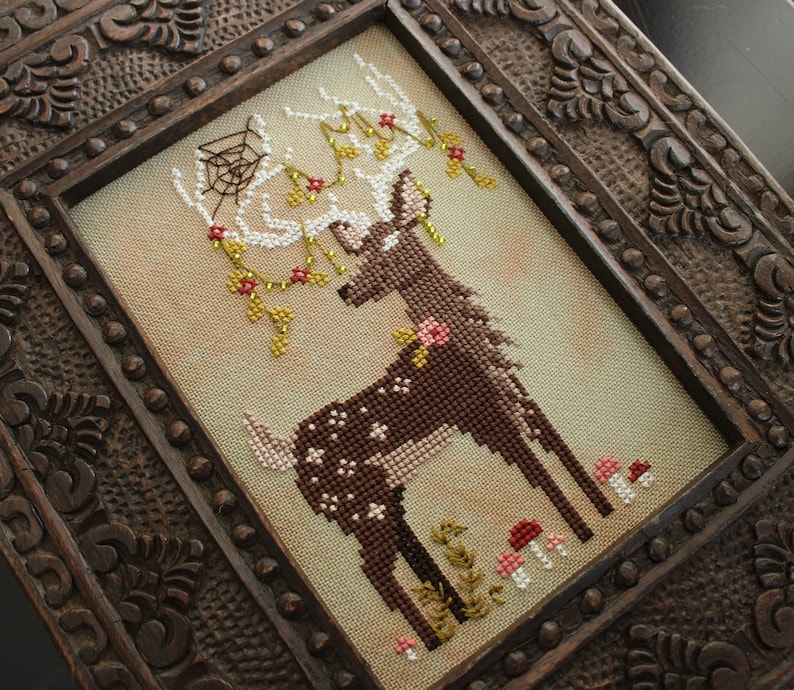 May include: Cross-stitch art of a deer with antlers adorned with a spiderweb, flowers, and berries. The deer is brown with white spots and is surrounded by mushrooms and foliage. The artwork is framed in a dark, intricately carved wooden frame.