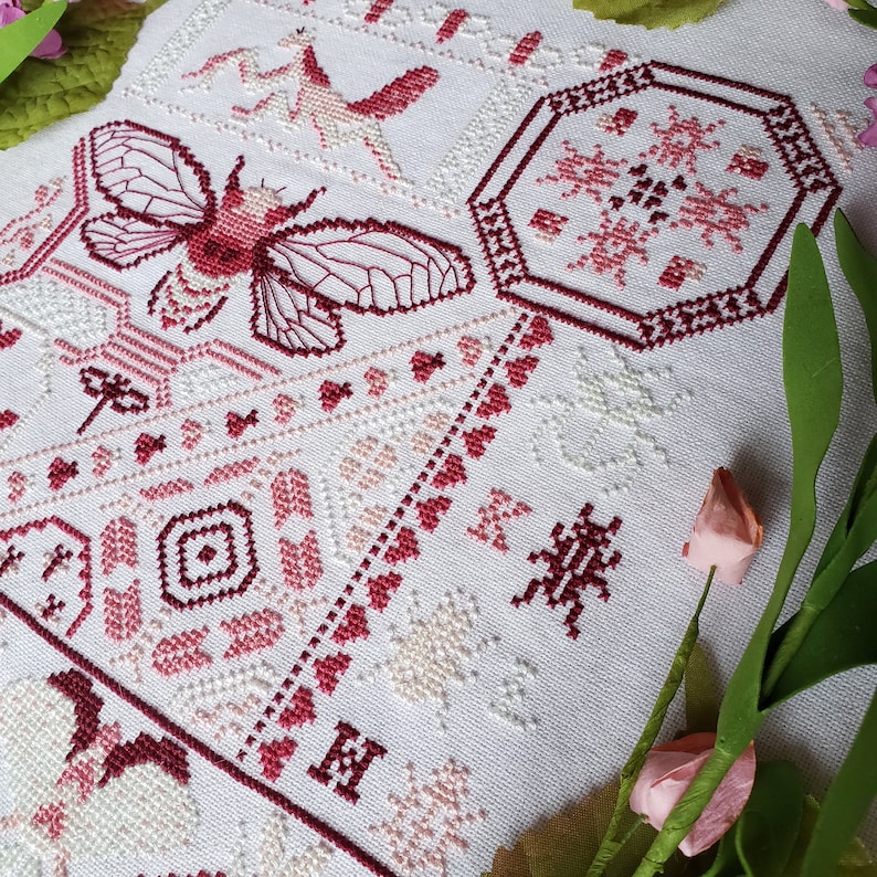 Another view of the finished pink cross stitch sampler of several insects and heart motifs, including a large cicada, praying mantis, and several moths.