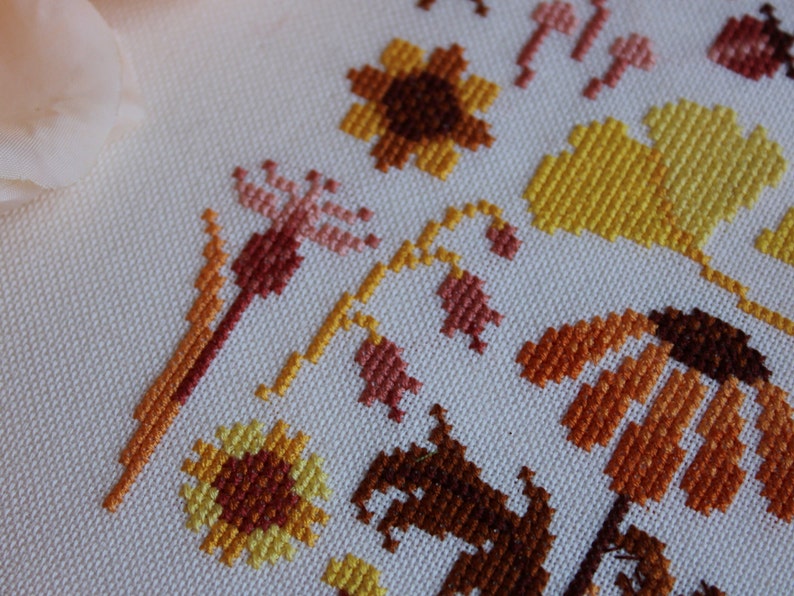 Peut inclure: Gros plan sur une broderie au point de croix repr&eacute;sentant une vari&eacute;t&eacute; de fleurs dans des tons de jaune, orange et marron. Les fleurs sont brod&eacute;es sur un fond de tissu blanc.