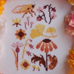 May include: Cross-stitch embroidery featuring a variety of autumnal flora and fauna, including a cicada, mushrooms, sunflowers, and ginkgo leaves in shades of yellow, orange, brown, and pink.