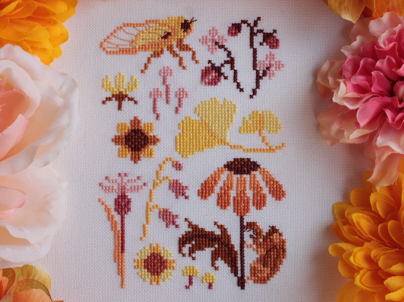 Peut inclure: Broderie au point de croix repr&eacute;sentant une vari&eacute;t&eacute; de flore et de faune automnales, notamment une cigale, des champignons, des tournesols et des feuilles de ginkgo dans des tons de jaune, orange, marron et rose.