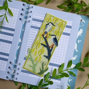 May include: A rectangular needlepoint bookmark featuring a cross-stitched design of two birds perched on a dark branch against a light yellow background with vertical green lines. The bookmark is placed on an open spiral-bound birding checklist.