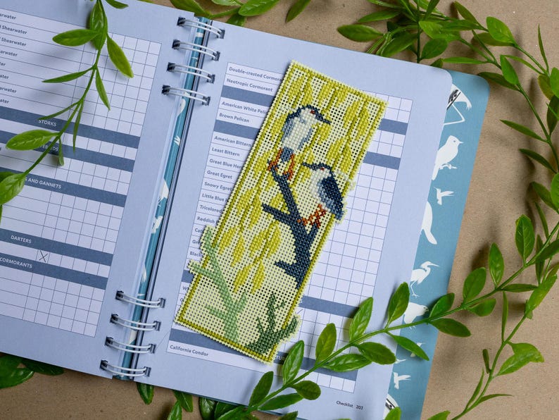 May include: A rectangular needlepoint bookmark featuring a cross-stitched design of two birds perched on a dark branch against a light yellow background with vertical green lines. The bookmark is placed on an open spiral-bound birding checklist.