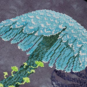 May include: Close-up of a cross-stitch design featuring a stylized blue and white wave. The wave is detailed with various shades of blue and white thread, set against a dark gray background. Green vines with small leaves are at the base of the wave.