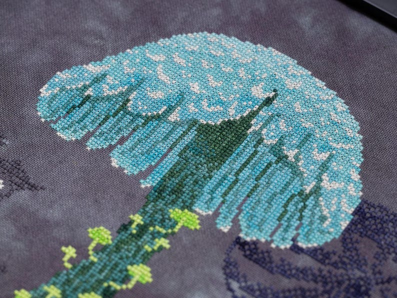 May include: Close-up of a cross-stitch design featuring a stylized blue and white wave. The wave is detailed with various shades of blue and white thread, set against a dark gray background. Green vines with small leaves are at the base of the wave.