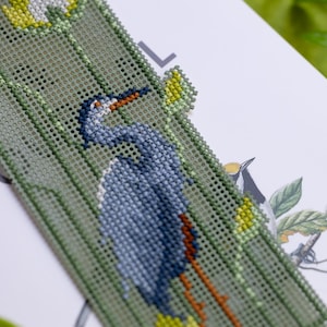 May include: A handmade bookmark featuring a detailed cross-stitch design of a blue heron with an orange beak and legs. The bookmark has a green background with yellow accents and is placed on a white surface.