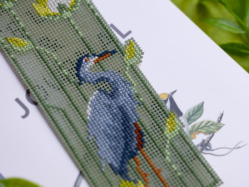 May include: A handmade bookmark featuring a detailed cross-stitch design of a blue heron with an orange beak and legs. The bookmark has a green background with yellow accents and is placed on a white surface.