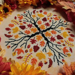 May include: A cross-stitch embroidery of a tree with autumn leaves in shades of red, orange, and yellow. The tree is surrounded by a border of fall-colored leaves.