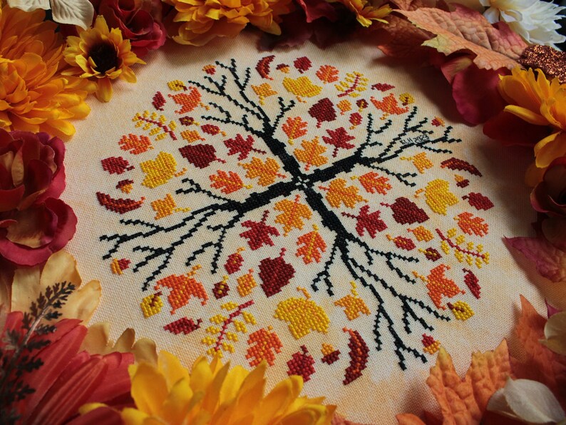 May include: A cross-stitch embroidery of a tree with autumn leaves in shades of red, orange, and yellow. The tree is surrounded by a border of fall-colored leaves.