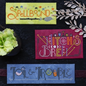 May include: Three cross-stitch samplers with Halloween themes. The top sampler is yellow with the word "SPELLBOUND" and a broom. The middle sampler is red with "WITCH'S BREW". The bottom sampler is blue with "TOIL & TROUBLE".
