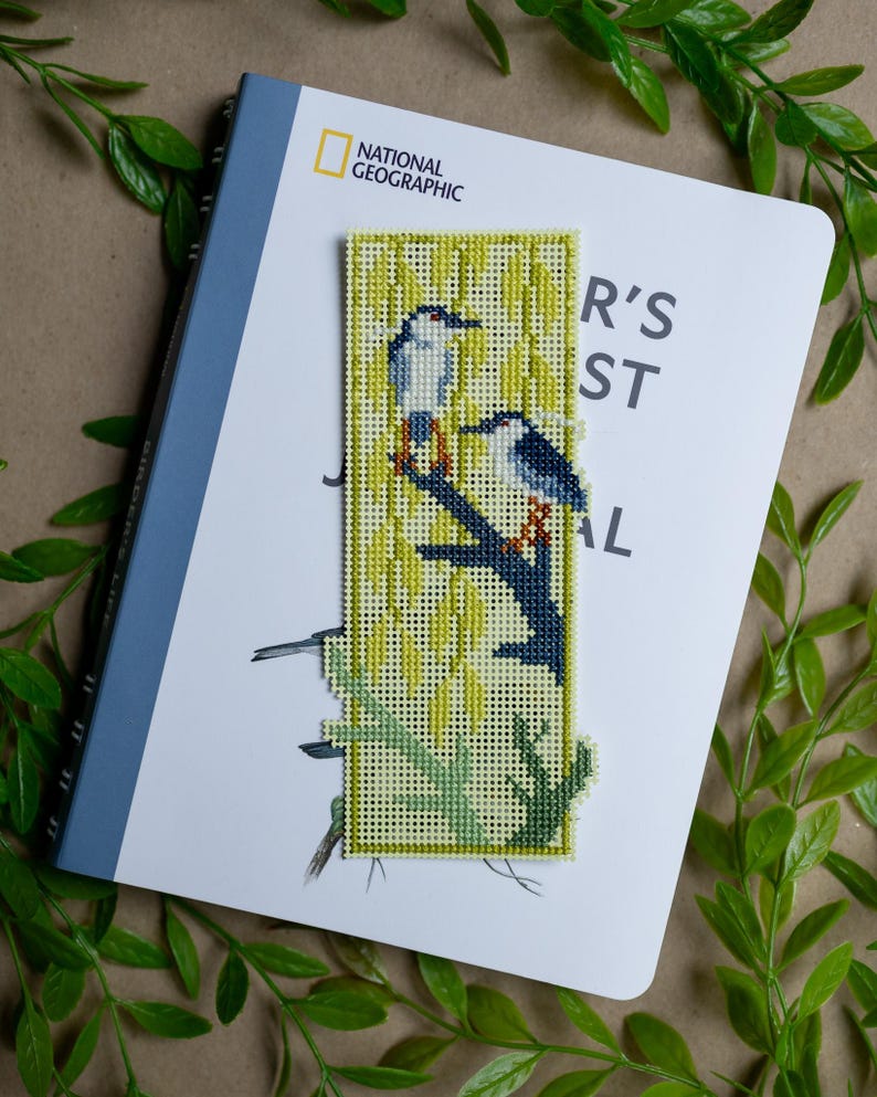 May include: A rectangular cross-stitch bookmark featuring two birds perched on a dark blue branch against a background of yellow willow leaves. The needlepoint design is stitched on a light yellow grid fabric with a distinct border and textured finish.