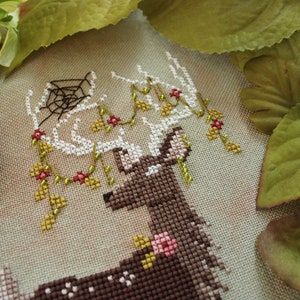 May include: Cross-stitch embroidery of a deer with antlers adorned with floral and spiderweb details. The deer is brown with white accents, and the flowers are pink and red. The background is a neutral beige.