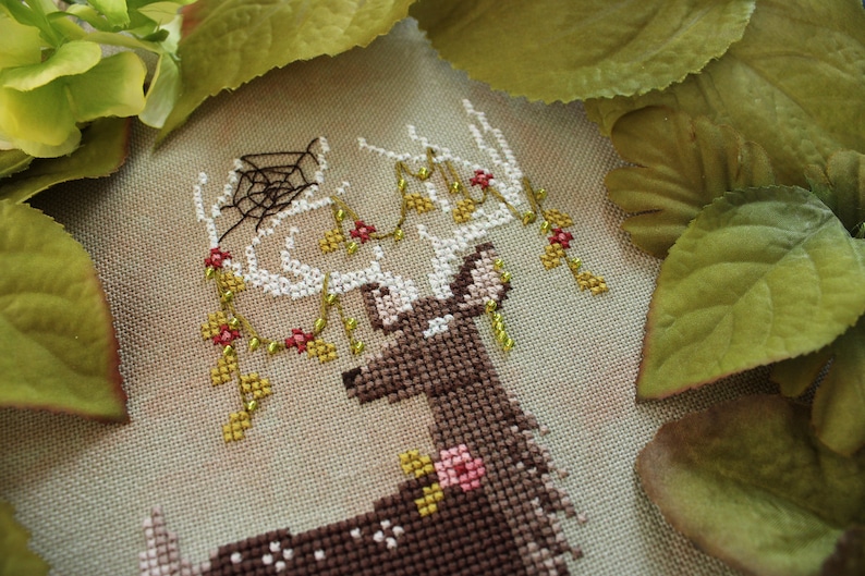 May include: Cross-stitch embroidery of a deer with antlers adorned with floral and spiderweb details. The deer is brown with white accents, and the flowers are pink and red. The background is a neutral beige.