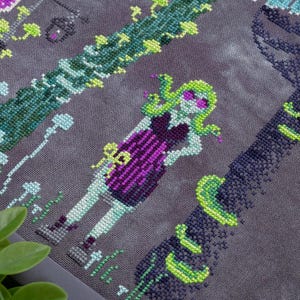 May include: Cross-stitch design featuring a stylized Medusa figure with green hair and sunglasses, wearing a purple dress. The design includes green vines and floral elements on a gray background. The artwork is detailed and colorful.