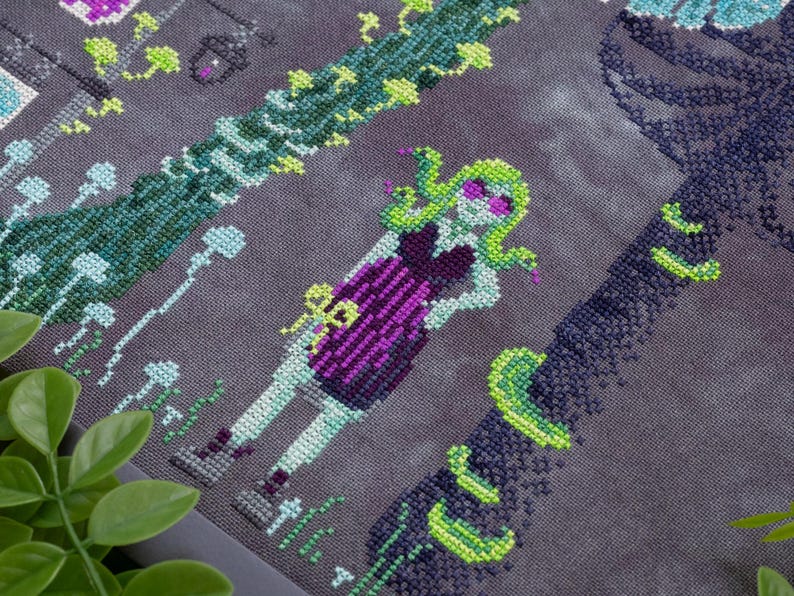 May include: Cross-stitch design featuring a stylized Medusa figure with green hair and sunglasses, wearing a purple dress. The design includes green vines and floral elements on a gray background. The artwork is detailed and colorful.