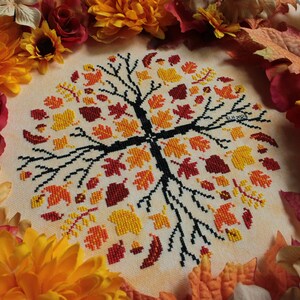 May include: A cross-stitch embroidery of a tree with autumn leaves in shades of red, orange, and yellow. The tree is surrounded by a border of fall-colored leaves and flowers.