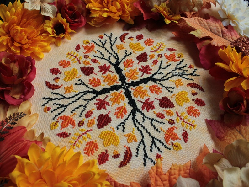 May include: A cross-stitch embroidery of a tree with autumn leaves in shades of red, orange, and yellow. The tree is surrounded by a border of fall-colored leaves and flowers.