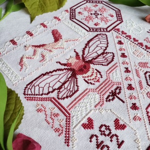 Another view of the finished pink cross stitch sampler of several insects and heart motifs, including a large cicada, praying mantis, and several moths.