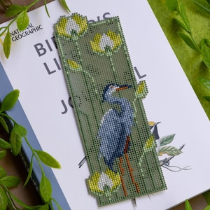 May include: A handmade bookmark featuring a cross-stitch design of a blue heron with yellow and white flowers on a green background. The bookmark is placed on a book with the text "Birds Life".
