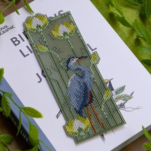 May include: A handmade bookmark featuring a blue heron design. The bookmark is made of green fabric with a cross-stitch pattern. The heron has blue and gray feathers, with a red beak and legs. Yellow and white flowers are also featured.