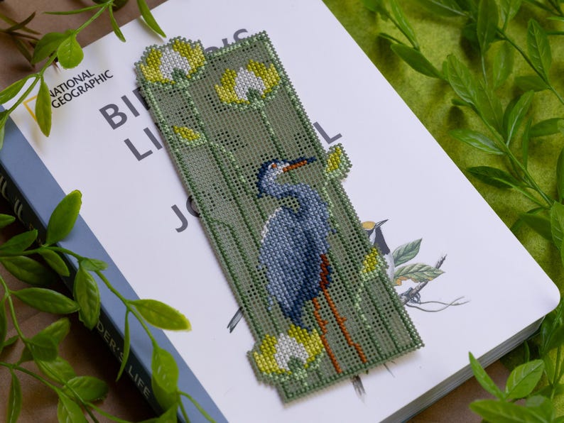 May include: A handmade bookmark featuring a blue heron design. The bookmark is made of green fabric with a cross-stitch pattern. The heron has blue and gray feathers, with a red beak and legs. Yellow and white flowers are also featured.